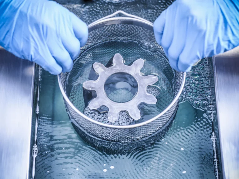 Två händer sänker ner en korg med en rund del i en ASONIC ultraljudsrengörare som är förberedd för rengöring. Händer som håller en trådkorg med ett stort kugghjul ovanför en ultraljudsrengörare som visar delarnas placering.